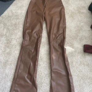 Bruna skinnbyxor från Divided, stl 36 - Snygga bruna byxor i fejkskinn från Divided (H&M) storlek 36. Byxorna har hög midja och raka ben med en lätt bootcut-känsla. Mjukt beige foder på insidan för extra komfort. Perfekta för dig som vill sticka ut med en trendig look.
