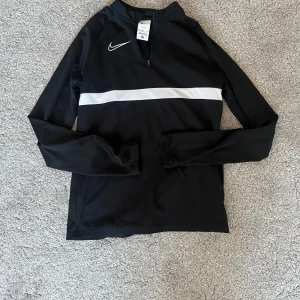 Nike svart träningströja Dri-Fit 158/170 - Svart långärmad träningströja från Nike med vit rand över bröstet och halv dragkedja vid halsen. Tillverkad i Dri-Fit material som andas och passar perfekt för sport eller träning. Snygg och sportig design med Nike-logga på bröstet.