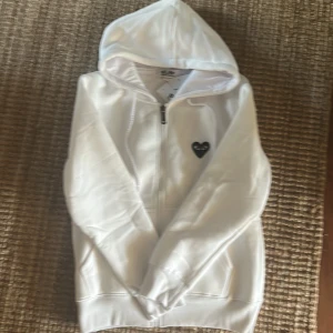 Vit hoodie Comme des Garçons Play S - Säljer en vit hoodie från Comme des Garçons Play i storlek S. Tröjan har dragkedja, huva med snörning och det ikoniska svarta hjärta-trycket med ögon både fram och bak. Tillverkad i mjuk bomull för en skön känsla.