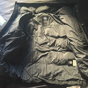 Svart  Canada Goose wyndam - Säljer en svart dunjacka från Canada Goose med klassisk patch på ärmen. Jackan har huva, dragkedja och tryckknappar framtill samt två stora fickor. Fodret är quiltat och jackan är perfekt för kalla dagar. Stilsäker och varm design med snygg passform.