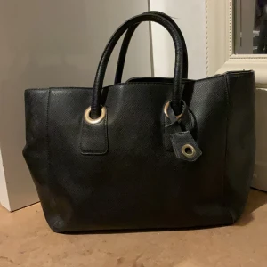 Svart handväska i skinn med detaljer - Stilren svart handväska i skinn med två rejäla handtag och snygga metalldetaljer vid fästena. Väskan har en klassisk, rymlig form och passar perfekt för dig som vill ha plats med allt du behöver. Enkel och tidlös design som funkar till många olika stilar.