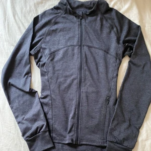 Mörkgrå träningsjacka från SOC - Sportig mörkgrå träningsjacka från SOC med hel dragkedja framtill och en ficka med dragkedja. Jackan är tillverkad i ett mjukt och stretchigt syntetmaterial som andas, perfekt för aktiva dagar. Långärmad modell med smal passform och diskret logga på ena sidan. Tröjan är i storlek 146-152 men sitter som en xxs-xs