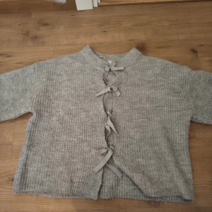Grå stickad kofta med snörning Gina Tricot - Mysig grå stickad kofta från Gina Tricot med snygg snörning framtill. Koftan har lång ärm, ribbad struktur och en rak, lite boxig passform. Perfekt för lager-på-lager under kyliga dagar.Har en förmörkning på enden på ena armen(se bild) använd ganska många gånger