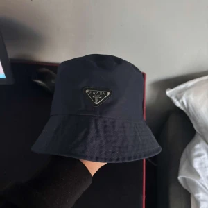 Prada mössa - Stilren svart bucket hat från Prada med klassisk form och ikonisk metallogo framtill. Hatten är tillverkad i ett slätt syntetmaterial som ger en modern look och passar perfekt för dig som vill ha en trendig accessoar med lyxig känsla. Nypris 6000