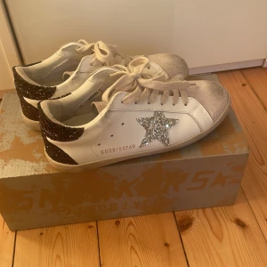 Golden Goose vita sneakers med glitter - Golden Goose sneakers i vitt skinn med silverglittrig stjärna på sidan och mörkbruna glitterdetaljer på hälen. Skorna är knappt använda och är i bra skick!