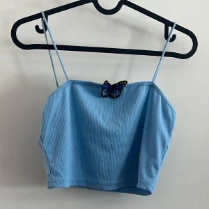 Ljusblå ribbad linne-topp  - Ljusblå ribbad linne-topp med tunna axelband och en broderad fjäril framtill som sticker ut. Linnetr en croppad modell 