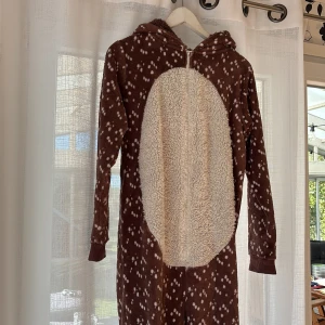 Brun jumpsuit med huva och djuransikte - Supermysig brun jumpsuit med huva, dekorerad med ett sött djuransikte och öron. Mjuk fleece med vita prickar och fluffig vit mage. Dragkedja framtill för enkel på- och avtagning. Perfekt för chill hemma eller roliga temakvällar.