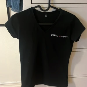 Svart v-ringad t-shirt Tommy Hilfiger - Marinblå t-shirt från Tommy Hilfiger med v-ringning och korta ärmar. Diskret logga broderad på bröstet i vitt, rött och blått. Skön passform och klassisk stil, perfekt till jeans eller shorts.