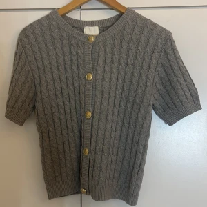 Grå kabelstickad kofta från H&M - Kortärmad grå kabelstickad kofta från H&M med snygga guldknappar framtill. Koftan har rund halsringning och ribbade muddar vid ärmslut och nederkant. Perfekt för lager-på-lager och en chill men ändå stilsäker look.