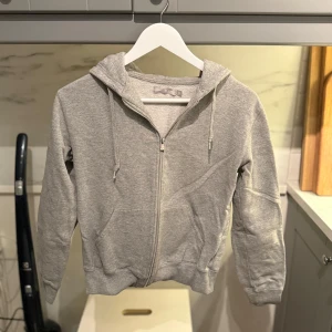 Grå hoodie med dragkedja och huva - Säljer en klassisk grå hoodie med dragkedja och huva. Tröjan har snörning i huvan, två fickor framtill och ribbade muddar vid ärmslut och nederkant. Den är från Soc.