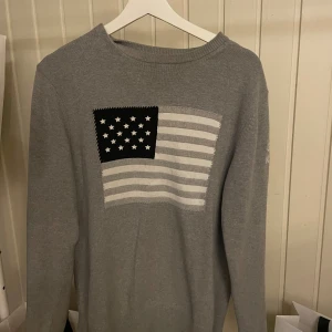 Grå stickad tröja från GANT - Snygg grå stickad tröja från GANT med amerikansk flagga i svart och vitt framtill. Tröjan har rund halsringning och långa ärmar, perfekt för dig som gillar klassisk men ändå cool stil.