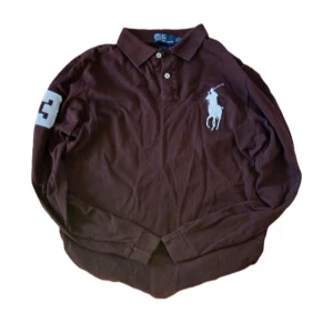 Brun långärmad pikétröja Ralph Lauren - Snygg brun långärmad pikétröja från Polo Ralph Lauren med klassisk krage, två knappar och stort vitt logobroderi på bröstet. Nummer 3 i vitt på ena ärmen. Tillverkad i mjuk bomull, perfekt för en chill och stilren look. Notera att en av knapparna har lossnat som du ser i bild 5. Skicka meddelande om du har någon fundering :)