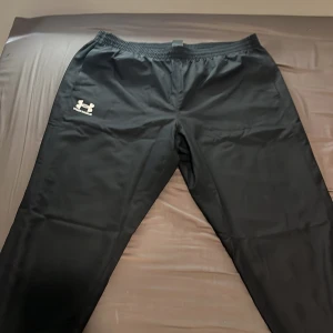 Svarta träningsbyxor Under Armour 2XL - Svarta träningsbyxor från Under Armour i storlek 2XL. Byxorna har elastisk midja, meshfoder och logga på vänster lår. Tillverkade i lätt polyester som andas, perfekta för gym eller sport. Snygg och enkel design med normal passform.