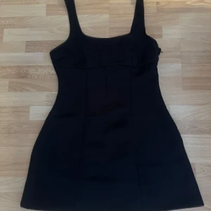 Ny. Svart kort klänning från H&M Divided - Supersnygg svart klänning från H&M Divided i storlek 38. Modellen är kort med breda axelband och figurnära passform. Materialet är slätt och känns som polyester, vilket ger en clean och modern look. Perfekt för dig som gillar minimalistisk stil.