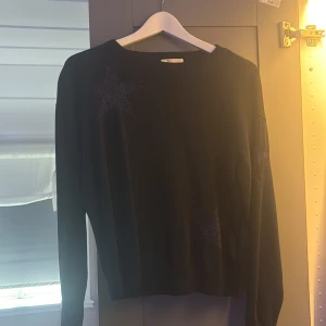 Svart stickad tröja med glitterstjärnor - Svart stickad tröja från Gina Tricot med glittriga stjärnor på bröstet, ärmen och nedtill. Rund hals och långärmad modell som är perfekt för dig som gillar lite extra detaljer. Mjuk och skön kvalitet som passar till många olika stilar.