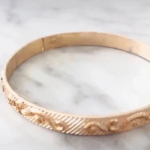 Guldigt armband med mönster - Snyggt guldigt armband med ett diskret, snirkligt mönster längs kanten. Armbandet har en rund form och passar perfekt för dig som gillar stilrena accessoarer med lite extra detalj. Perfekt att kombinera med andra smycken för en personlig look.