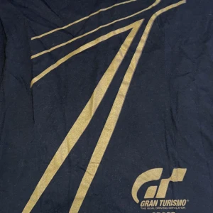 Gran Turismo svart t-shirt Gildan - Svart t-shirt från Gildan med stort guldtryck och Gran Turismo-logo framtill. Klassisk passform med korta ärmar och rund hals. Tillverkad i mjuk bomull, perfekt för dig som gillar gaming och snygga prints.
