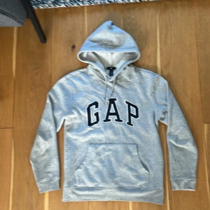 Grå hoodie från GAP - Säljer en klassisk grå hoodie från GAP med stor svartvit logga framtill. Tröjan har huva med snörning, magficka och är tillverkad i mjuk bomullsblandning. Perfekt för chill dagar eller när du vill ha en enkel och snygg look liten fläk/defekt på armen fråga gärna om bild.