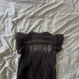 Svart t-shirt med volangärmar Gina Tricot - Svart t-shirt från Gina Tricot med coola volangärmar och ribbad struktur. Framsidan har texten 'YOUNG' i glittriga paljetter som ger extra attityd. Perfekt för dig som gillar statement-plagg och vill sticka ut lite extra.