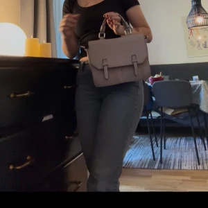 Beige axelväska med spänndetaljer - Säljer en stilren beige axelväska i skinnimitation med snygga spänndetaljer framtill. Väskan har både handtag och justerbar axelrem, vilket gör den enkel att bära på olika sätt. Perfekt storlek för det viktigaste och passar till många outfits. 1 dragkedefack och 2 andra mindes fack i väskan