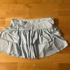 Ljusblå volangkjol med bred midja - Supersöt ljusblå kortkjol med dubbla volanger och bred midja. Kjolen har en flowy känsla och är perfekt för dig som gillar en feminin och lekfull stil. Tillverkad i mjukt bomullstyg som känns skönt mot huden. Med shorts under