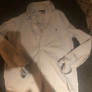 Ljusblå skjorta från Polo Ralph Lauren - Snygg ljusblå skjorta från Polo Ralph Lauren i slim fit-modell. Klassisk button-down krage, långa ärmar och broderad logga på bröstet. Tillverkad i mjuk bomull och stängs med vita knappar framtill. Perfekt för en clean och stilren look. Kan gå ner i pris vid snabbt köp