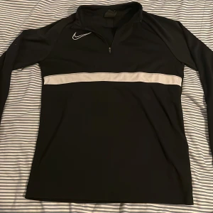 Nike svart half zip träningströja L - Svart långärmad träningströja från Nike i storlek L men passar S. rand över bröstet och diskret Nike-logga. Tröjan har half zip och är gjord i Dri-Fit polyester som andas. Perfekt för sport eller chill, stilren och skön passform.