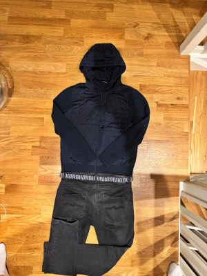 Mörkblå hybridjacka från J.Lindeberg - Svart Pierre Knitted Hybrid Jacket från J.Lindeberg med quiltad front och rygg i syntet, stickade ärmar och huva. Jackan har dragkedja framtill och en diskret logga på bröstet. Perfekt för dig som gillar stilren och sportig look. Perfekt nu inför vintern och om du har några funderingar är de bara att höra av sig🫡