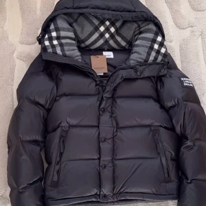 Svart pufferjacka från Burberry - Säljer en svart pufferjacka från Burberry med klassiskt rutigt foder i huvan. Jackan har dragkedja och knappar framtill, två sidofickor med dragkedja och en snygg patch på ärmen. Perfekt för kalla dagar och riktigt stilren look.