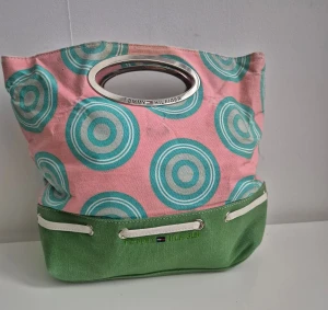 Mönstrad handväska Tommy Hilfiger - Snygg handväska från Tommy Hilfiger med rosa och grönt tyg, coola turkosa cirkelmönster och metallhandtag. Väskan har grön botten med vita detaljer och insidan är randig i blått och vitt med dragkedja. Perfekt för dig som gillar färg och mönster. OBS ... små skavanker dvs missfärgningar 