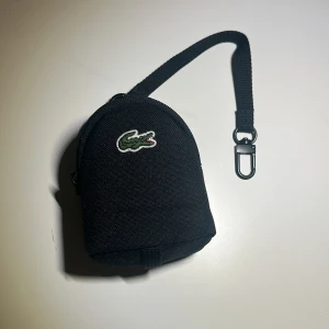 Svart myntväska från Lacoste - Liten svart myntväska från Lacoste med klassisk krokodillogga framtill. Väskan har en rundad form, dragkedja och en praktisk rem med karbinhake. Tillverkad i slitstark polyester, perfekt för småprylar och coins eller kort.