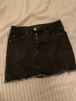 Svart jeanskjol från American Eagle - Svart jeanskjol från American Eagle med rå kant nertill och klassisk femficksdesign. Kjolen har hög midja, bälteshällor och knäppning framtill. Lite ljusare svart i färgen, är i storlek Uk 2 men passar mig som har storlek 34. Är  i använt men bra skick och säljs i befintlig skick. 