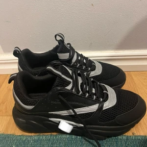 Svarta sneakers från Italien, strl 44 - Snygga svarta sneakers med silvriga detaljer och mesh-paneler. Skorna har rund tå, platt sula och snörning framtill. Tillverkade i Italien, storlek 44. Perfekt för dig som gillar sportig och modern stil med coola kontraster.