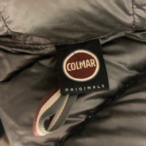 Svart Colmar pufferjacka - Säljer en svart/mörkblå pufferjacka från Colmar Originals med klassisk logga på ärmen. Jackan har en rak passform, långa ärmar och grova horisontella sömmar. Dragkedja framtill och lätt glansigt material som ger en snygg look. Perfekt för kyliga dagar. (Finns defekt på ena ärmen, sista bilden. Men syns knappt när den används och går lätt att sy!)