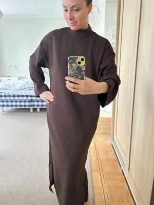 Brun långklänning med slits - Säljer en stilren brun långklänning med rak passform och höga slitsar på båda sidor. Klänningen har lång ärm och en hög krage som ger en cool och minimalistisk vibe. Perfekt för dig som gillar enkla och snygga plagg med lite edge.