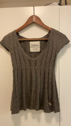 Grå stickad topp från Abercrombie - Säljer en grå stickad topp från Abercrombie med korta ärmar och v-ringning. Toppen har ett snyggt kabelstickat mönster och små håldetaljer längs fram och bak. Mjuk och skön, perfekt för lager-på-lager. Liten logga broderad nedtill.
