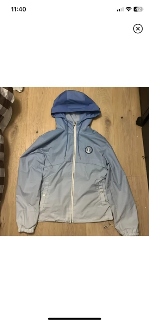 Blå vindjacka med huva och dragkedja - Snygg vindjacka i två nyanser av blått med huva och vit dragkedja framtill. Jackan har ett emblem på bröstet, snörning i nederkant och praktiska fickor med knappstängning. Perfekt för dig som gillar sportig och clean stil.