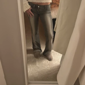 Grå bootcut jeans med låg midja - Snygga grå jeans med bootcut och låg midja. Jeansen har klassisk femficksdesign, silverfärgade knappar och dragkedja. Materialet är jeans med en lätt stretchig känsla och tvättad look. Perfekt passform för dig som gillar retrovibbar och markerad siluett.