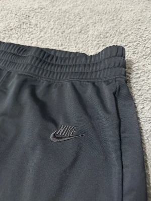Svarta träningsbyxor från Nike - Nike träningsbyxor i svart med broderad logga på benet. De har elastisk midja och är tillverkade i ett mjukt, syntetiskt material som är perfekt för sport och chill. Slim passform och stilren design gör dem enkla att matcha med din favorittröja.
