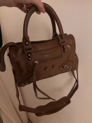 Brun handväska med dragkedja - Snygg brun handväska i skinn med coola metalldetaljer och dragkedja framtill. Väskan har både handtag och axelrem, vilket gör den enkel att bära på olika sätt. Perfekt storlek för det viktigaste och har en trendig look med dekorativa nitar.
