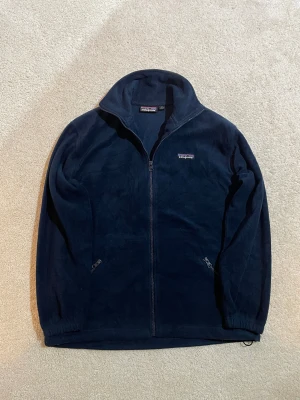 Mörkblå fleecejacka från Patagonia | M - Mysig mörkblå fleecejacka från Patagonia med hel dragkedja och högt krage. Jackan har två sidofickor med dragkedja och justerbar nederkant med snörning. Perfekt för kyliga dagar och outdoor-häng. Diskret Patagonia-logga på bröstet.