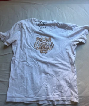 Vit Kenzo t-shirt med tigertryck - Säljer en vit t-shirt från Kenzo i bomull med ett snyggt guldigt tigertryck på bröstet. Klassisk rund hals och korta ärmar. Perfekt för dig som gillar streetstyle och vill sticka ut med ett ikoniskt motiv.
