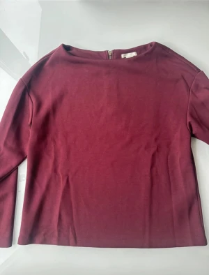 Vinröd långärmad topp med dragkedja - Vinröd långärmad topp med rund hals. Toppen har en snygg synlig dragkedja i ryggen som ger en cool detalj. Perfekt att styla med jeans eller kjol för en chill look