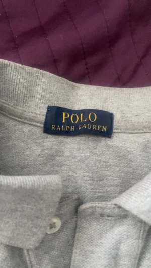 Grå pikétröja från Polo Ralph Lauren - Klassisk grå pikétröja från Polo Ralph Lauren med broderad logga på bröstet. Tröjan har krage, två knappar och är tillverkad i mjuk bomull. Perfekt för en clean och stilren look.