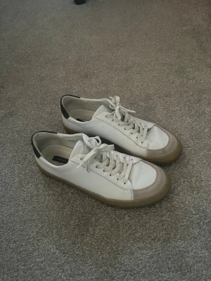 Vita sneakers från Sneaky Steve - Snygga vita sneakers från Sneaky Steve med beige sula och svart detalj på hälen. Skorna har klassisk snörning och är gjorda i skinn. Perfekta för dig som gillar en clean och stilren look. Nypris: 1900kr