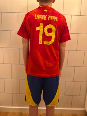 Spaniens Lamine Yamal matchtröja #19 - Säljer en officiell Spanien fotbollströja med Lamine Yamal och nummer 19. Tröjan är röd med gula detaljer och tryck, kortärmad och tillverkad i lätt polyester. Landslagsemblem på bröstet och matchande blå shorts med gula inslag och nummer 19.