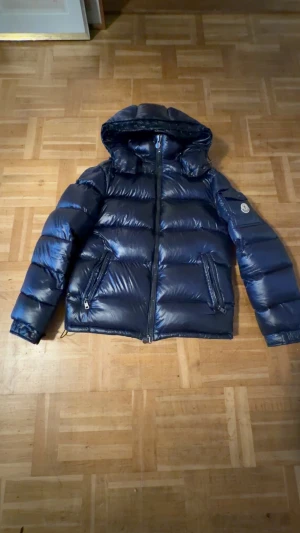 Moncler Maya - Säljer en mörkblå pufferjacka från Moncler med glansig finish, huva och två dragkedjeförsedda fickor. Jackan har brunt foder med en cool serietidningsetikett på insidan. Perfekt för kalla dagar och har en klassisk Moncler-logga på ärmen.     Storlek 1 är lika med storlek S