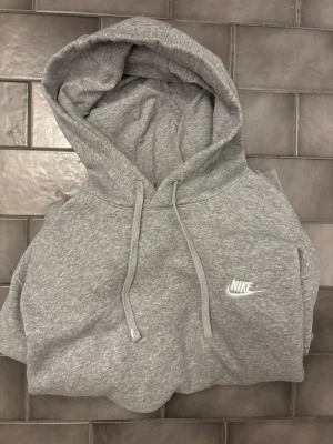 Grå hoodie från Nike med huva - Säljer en grå hoodie från Nike med klassisk logga broderad i vitt på bröstet. Tröjan har huva med snörning och en avslappnad passform. Perfekt för chill dagar eller när du vill ha en enkel och snygg look.