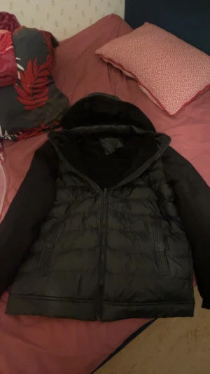 Svart pufferjacka med huva - Svart pufferjacka med huva och quiltad design. Jackan har långa ärmar i slät polyester och två fickor med snygga ribbade detaljer och knapp. Dragkedja framtill för enkel stängning. Perfekt för kalla dagar när du vill hålla stilen.