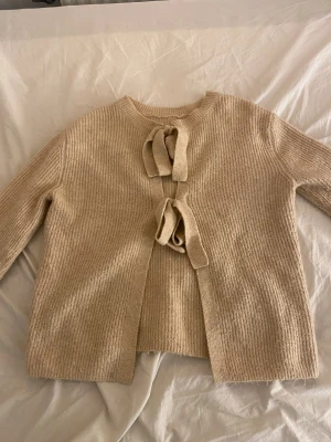 Beige stickad kofta med knyt från Only - Säljer en beige stickad kofta med två snygga knytband framtill. Koftan har ribbad struktur och långärmade ärmar, perfekt för att ge en chill och mysig vibe. Passar dig som gillar neutrala färger och enkel stil.